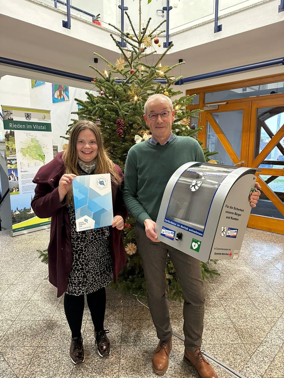 Wirtschaftsförderin Angela Seidel und Bgm. Erwin Geitner bei der AED-Bescheidübergabe an die Gemeinde Rieden vor einem Weihnachtsbaum 